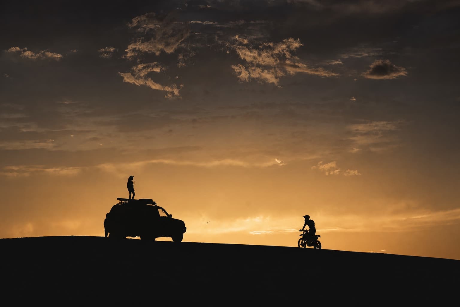 A field vehicle, rider and small group silhouetted together on a dune at sunset