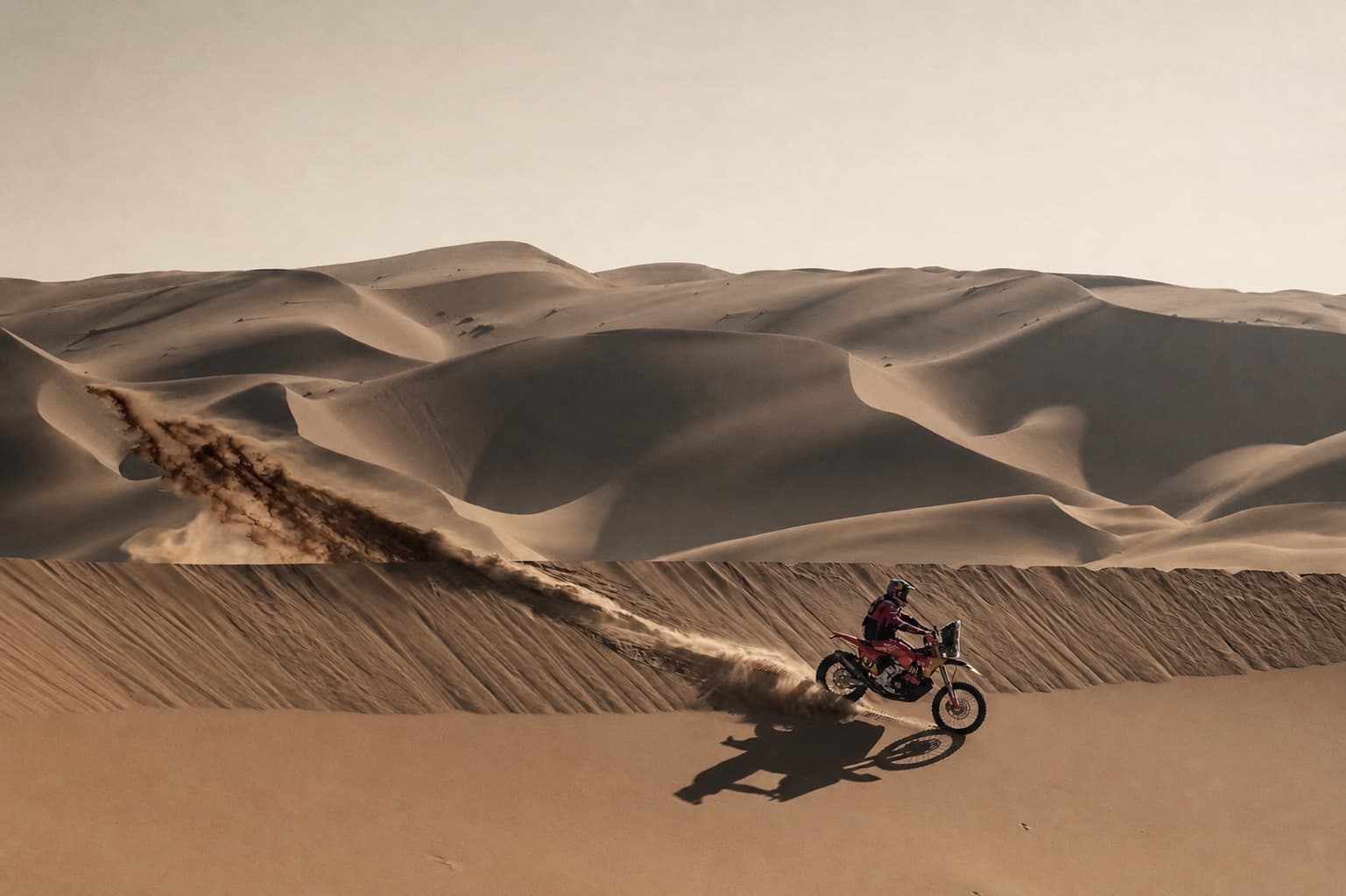 A lone motorcyclist following a long sand trail across layered dunes